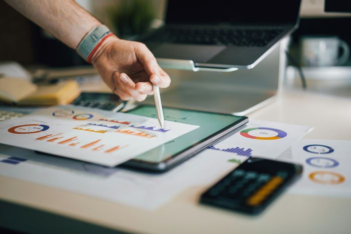 Auf dem Schreibtisch befinden sich ein Laptop, ein Tablet, ein Taschenrechner und Blätter mit einer Präsentation verschiedener Daten. Die Hand eines Mannes deutet mit einem Stift auf einige Diagramme